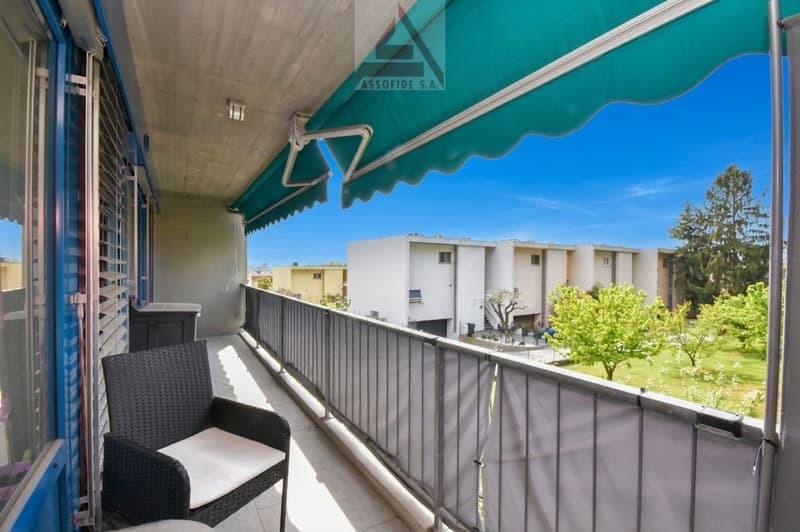 Balcony with metal railing, green awning, black wicker chair, white cushion, metal storage box, view of a garden and buildings