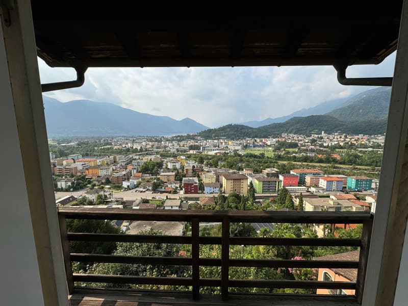 The image shows a panoramic view of a city surrounded by mountains. The view is seen through a balcony or terrace, with a wooden railing in the foreground. The city appears to be a densely populated urban area with a mix of residential and commercial buil