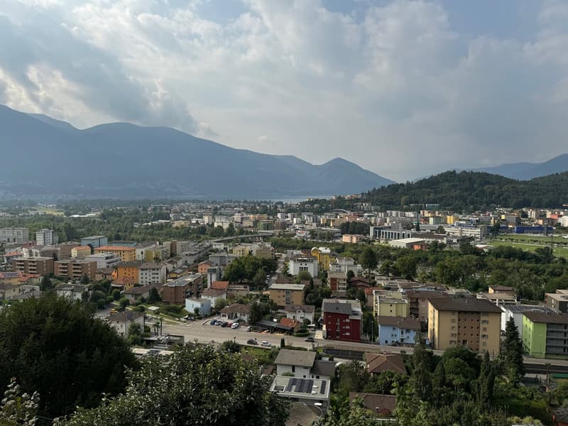 The image shows a panoramic view of a city surrounded by mountains. The city has a mix of residential and commercial buildings, with some high-rise structures visible. The landscape is dotted with trees and greenery, and the sky is partly cloudy. The over