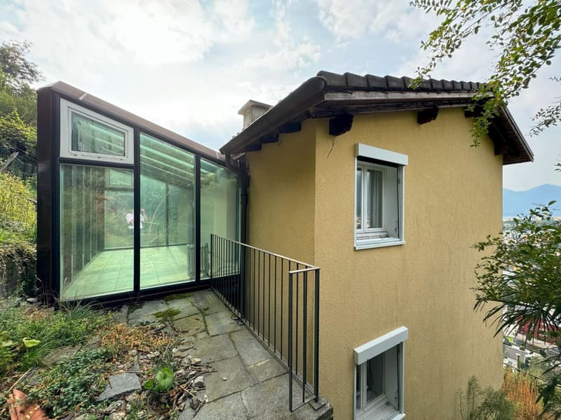 Single-story house with yellow exterior walls, large glass windows, and a balcony or terrace with a metal railing. The house has a slanted roof and is surrounded by greenery.