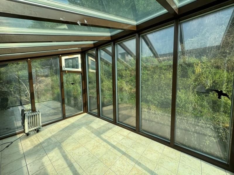 Large glass-enclosed sunroom with floor-to-ceiling windows, providing a panoramic view of the surrounding nature. The room has a tiled floor and a radiator, suggesting it is a comfortable and well-insulated space.