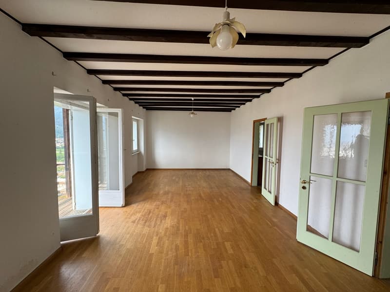 Large open room with hardwood floors, exposed wooden beams on the ceiling, and French doors leading to a balcony or terrace. The room has a clean, minimalist design with white walls and green-framed doors.