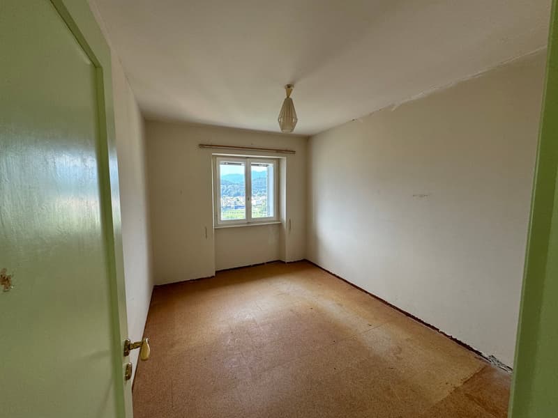 The image shows a simple, empty bedroom with white walls, a window overlooking a view, and a light fixture on the ceiling. The room appears to have a hardwood or linoleum floor.