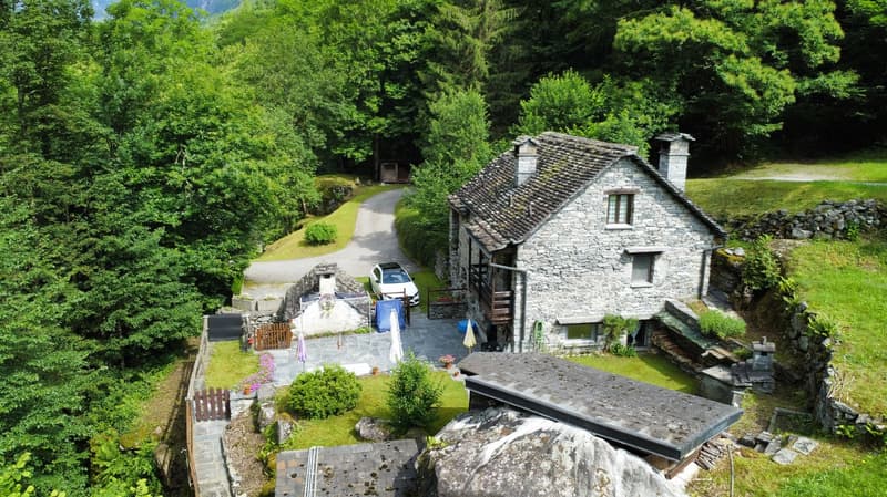 Stone house, two chimneys, car parked in front, surrounded by trees and grass, driveway leading to the house