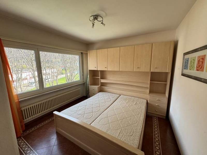 Bedroom with wooden bed, wooden cabinets, large window, radiator, ceiling lamp, and a picture frame on the wall