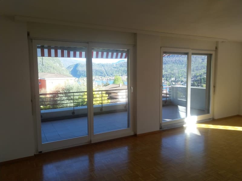 An empty room with large sliding glass doors opening onto a balcony with a view of mountains and a lake.
