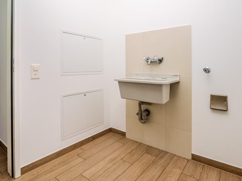 The image shows a utility room with a white wall, a built-in cabinet, and a sink with a faucet. The floor is made of wood-like tiles.
