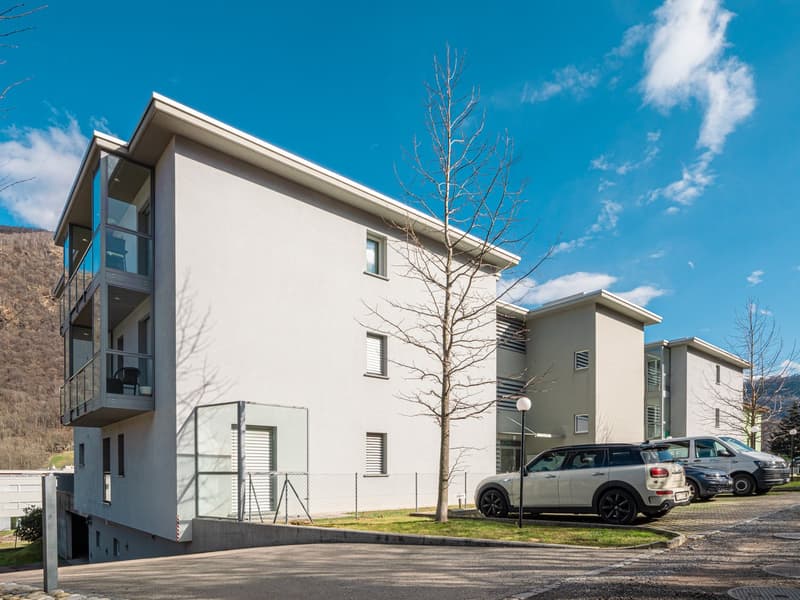 3-story modern apartment building with white exterior, large windows, and balconies. There is a parking lot in front of the building with several vehicles parked.