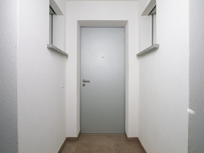 The image shows a simple, minimalist entrance area with a white door, white walls, and a tiled floor. There are two small shelves or ledges on either side of the doorway, likely for storage or decoration. The overall aesthetic is clean and modern.