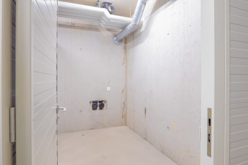 This image shows a small, unfinished room or storage space. The walls are bare and unfinished, with exposed electrical outlets and ventilation pipes. The floor appears to be a concrete or cement surface. The room has a simple door and seems to be used for