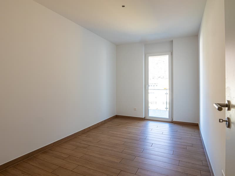 This image shows an empty room with white walls and a hardwood floor. The room has a single window that provides natural light. The room appears to be a bedroom or living space, with no visible furniture or other features.