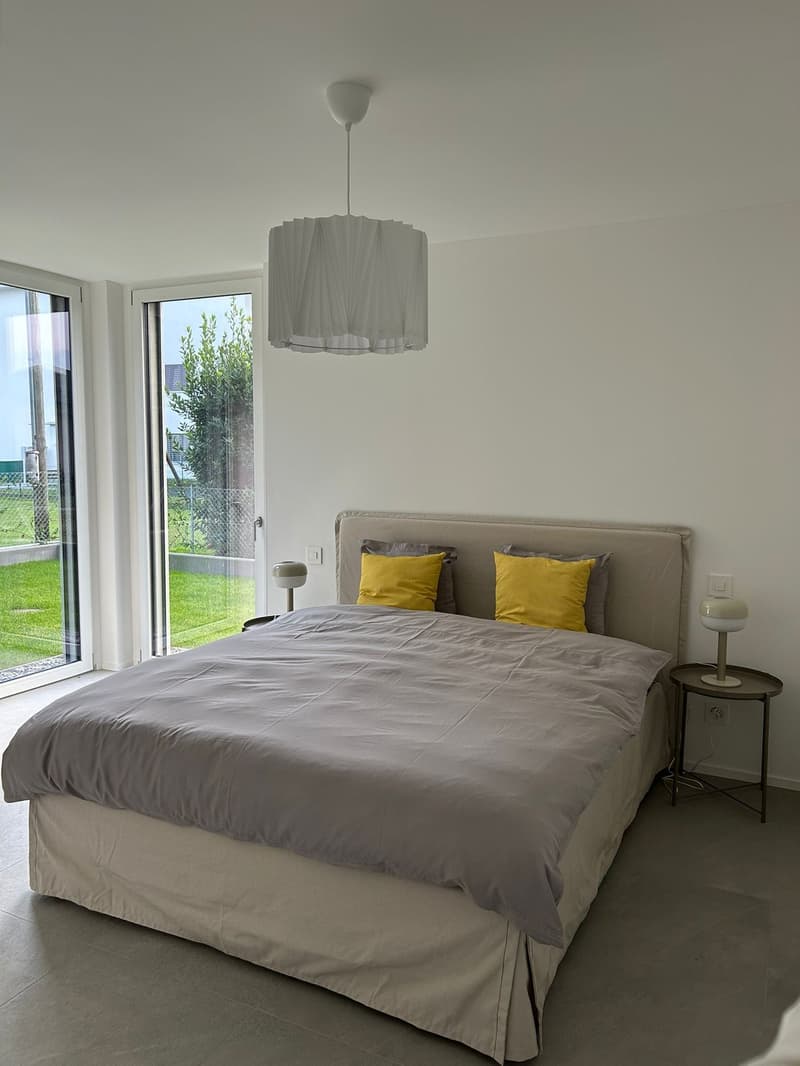modern bedroom, large bed, gray bedspread, white ceiling lamp, white walls, double glass doors, large windows, view to the garden, wooden bedside tables, yellow pillows