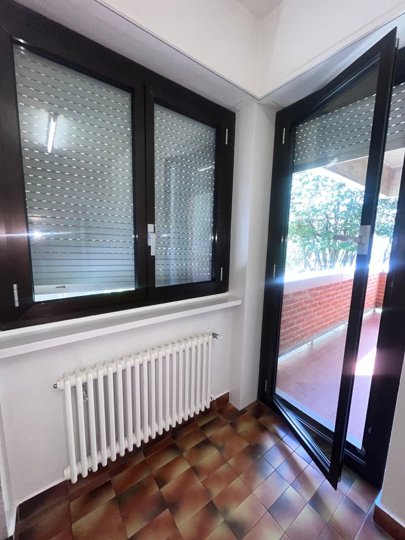 Empty room with white radiator, tiled floor, two windows with blinds, open door to balcony