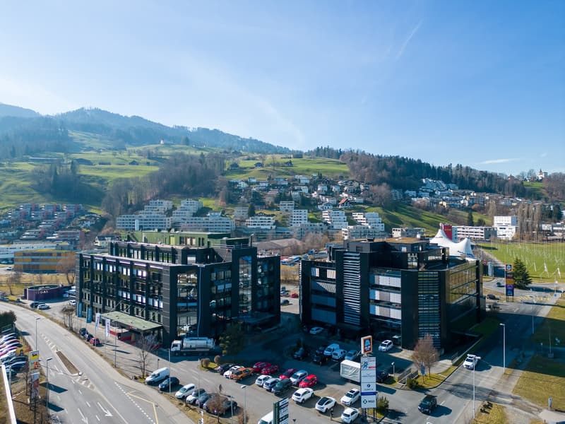 Aerial view of a commercial building complex with multiple floors, surrounded by a parking lot and other commercial buildings, located in a mountainous region with a lush landscape.