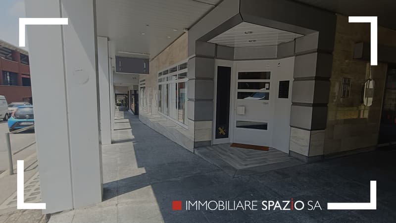 Exterior of a commercial building with tiled flooring, white exterior walls, a large entrance door, and signage indicating 'Immobiliare SPAZIO SA'. There are some cars parked on the street and buildings visible in the background.