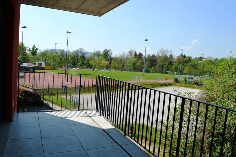 A balcony with black railing, a tiled floor, overlooking a grass field with an athletic track, mountains in the distance