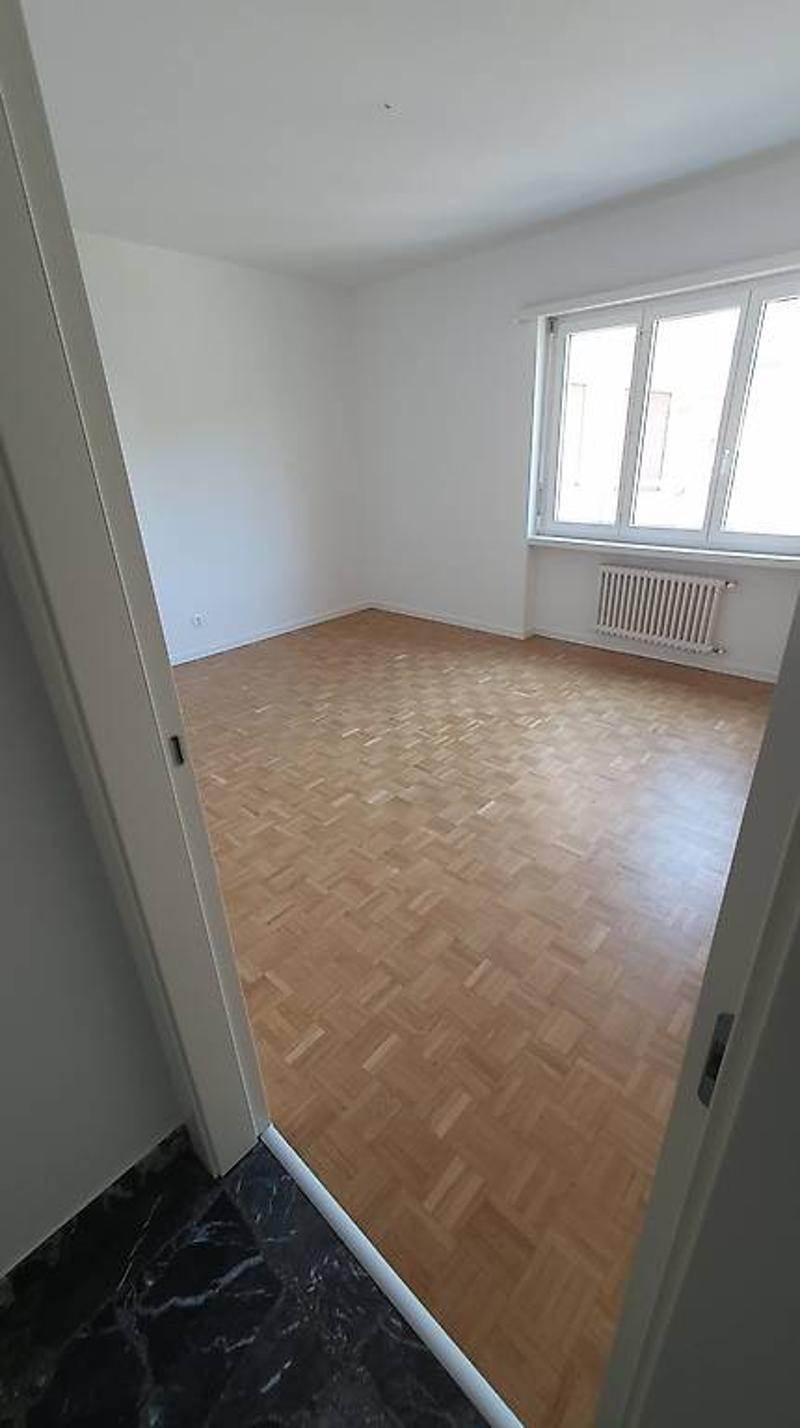 Empty room with white walls, hardwood parquet flooring, and a window.