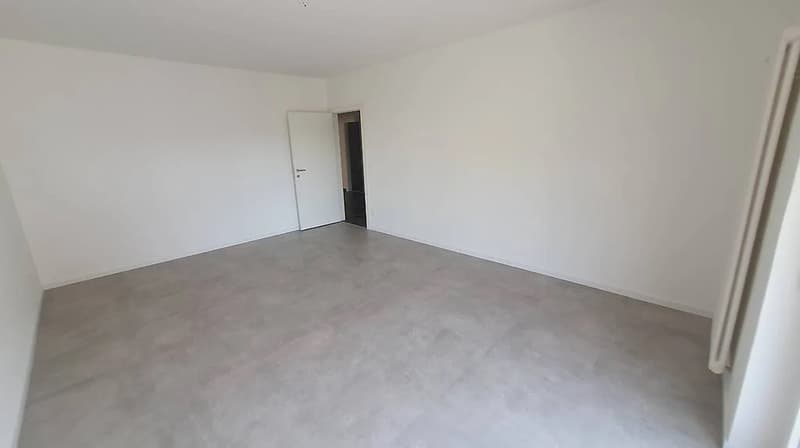 Empty room with white walls and gray concrete flooring. The room has a door visible in the background.