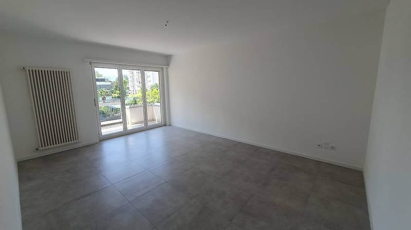 Spacious living room with large windows, sliding glass doors leading to a balcony or terrace, and gray tile flooring.