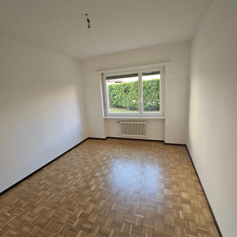 Empty bedroom with white walls, hardwood parquet flooring, and a large window overlooking greenery outside.