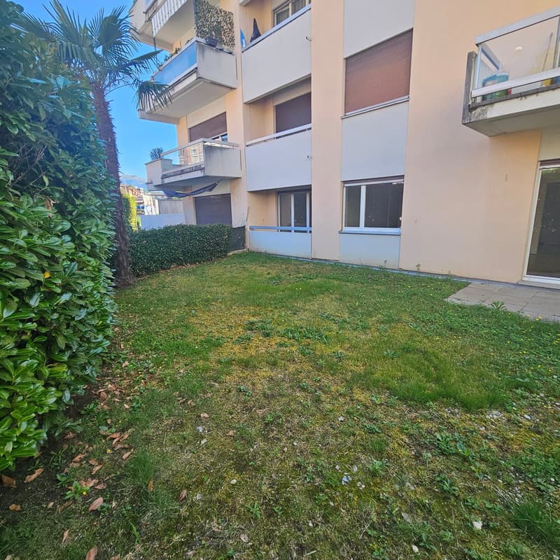 Multi-story apartment building with balconies, surrounded by greenery including palm trees and a grassy area in the foreground.