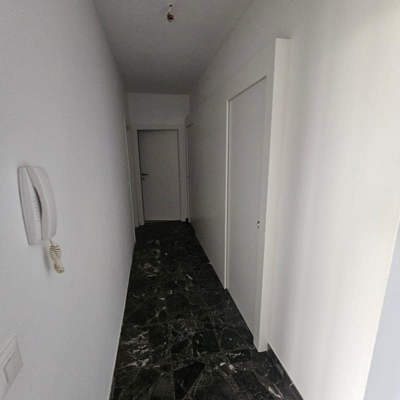 Long, narrow hallway with white walls and a black marble-patterned floor. There is a white door at the end of the hallway and a white intercom system on the wall.