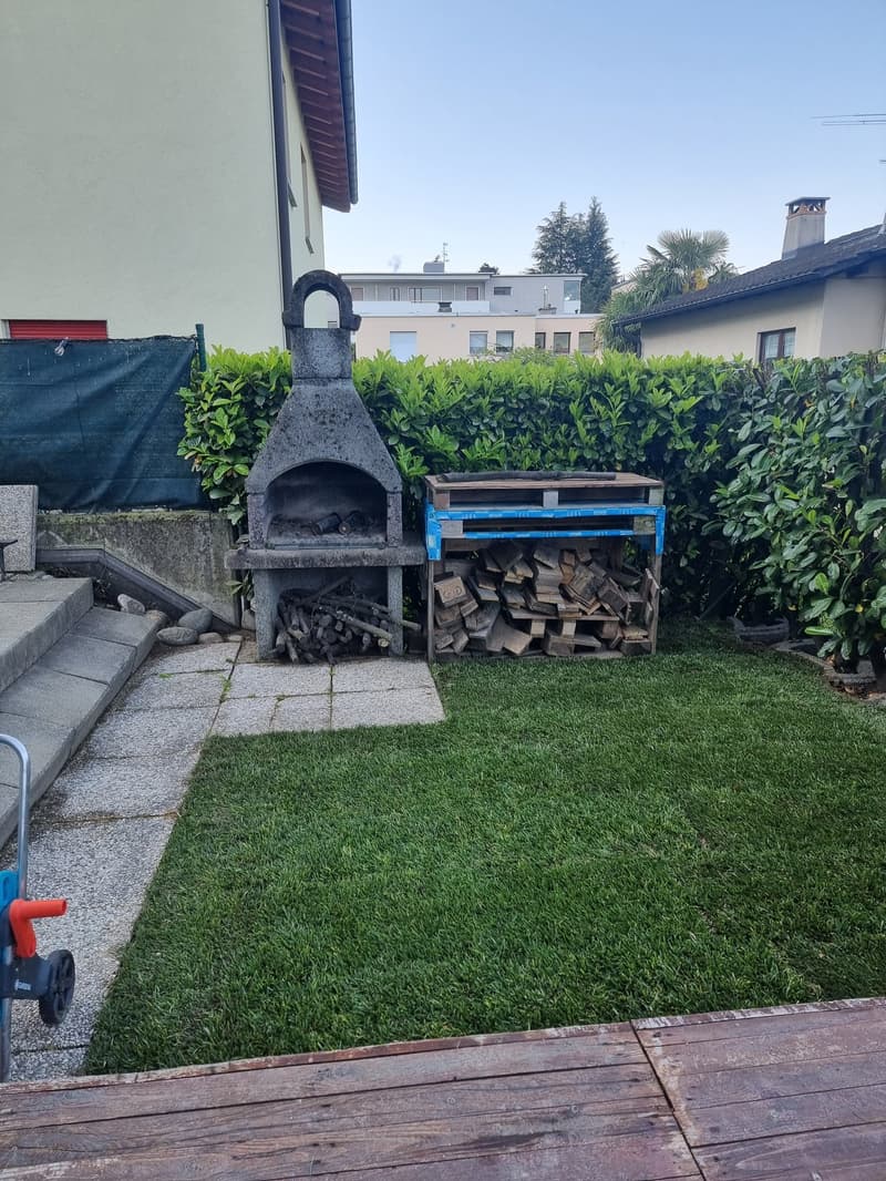 Outdoor fireplace, wood storage, green lawn, wooden deck, steps, hedges, and a building in the background.