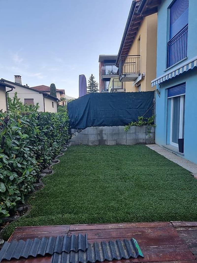 A backyard with a wooden deck, a green lawn, and a concrete wall with a black cover.
