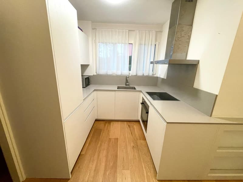 Modern L-shaped kitchen with white cabinets, wooden floors, stainless steel appliances, and ample natural light from the window.