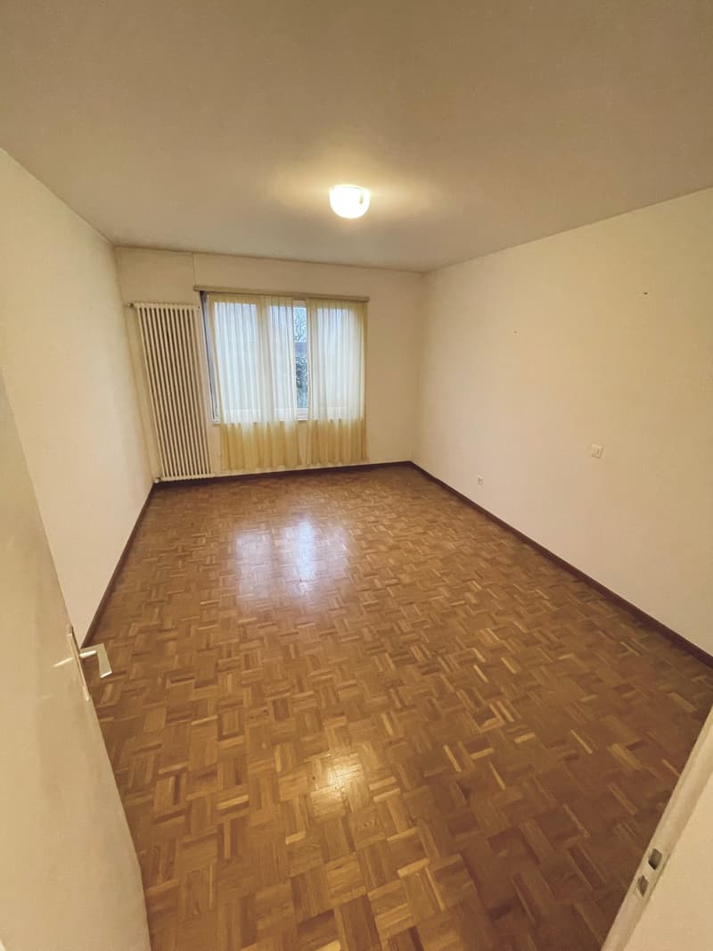 large room with wooden parquet flooring, white walls, large window with white curtains, white radiator