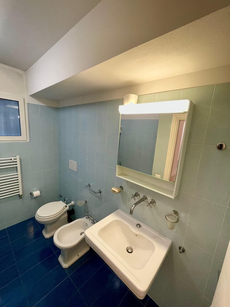Tiled walls, tiled floor, white sink, silver faucet, mirror with lights, bidet, toilet, towel rack, window