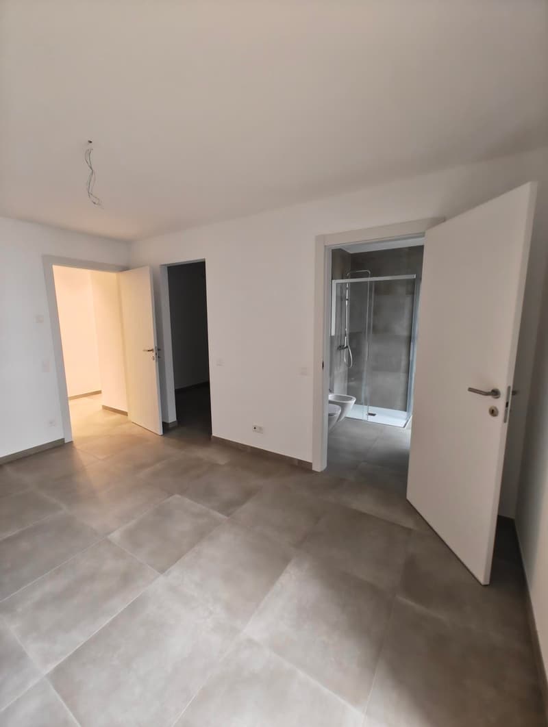 An empty room with white walls and tiled floor. There is an open door leading to another room. There is a glass shower room with a toilet visible through the open door.