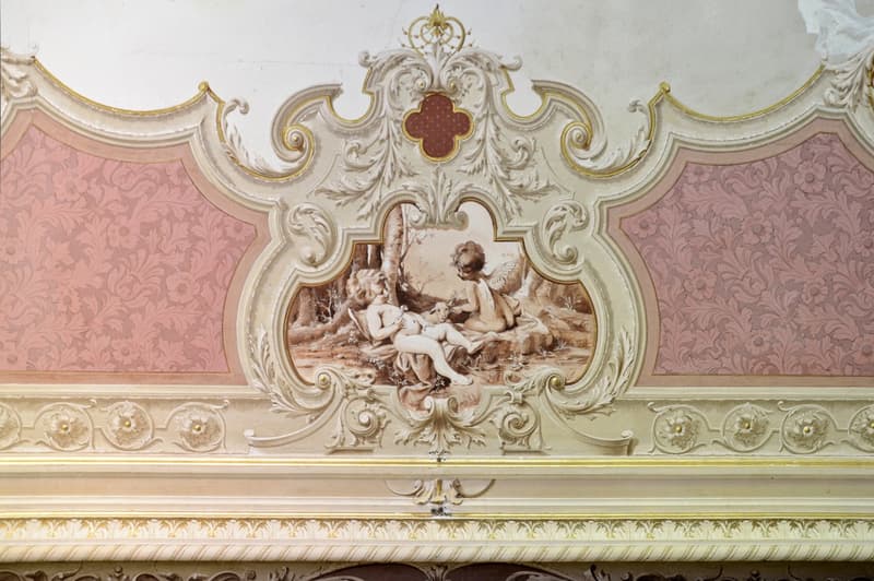 The image shows an ornate, baroque-style architectural detail featuring intricate carvings, gilded accents, and a central painting depicting a mythological scene with cherubs and figures. The overall design is highly decorative and ornamental, with a pink