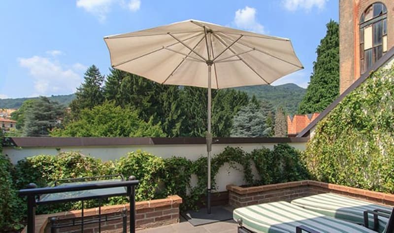 The image shows an outdoor patio or terrace area with a large beige umbrella, a bench, and a table. The patio is surrounded by greenery, including trees and shrubs, and there appears to be a parking area or driveway visible in the background.
