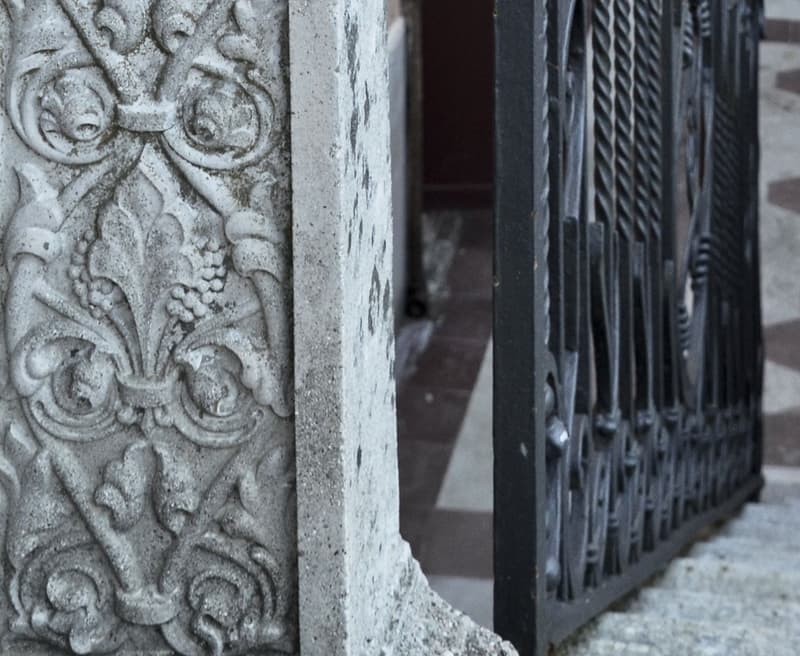 The image shows a close-up view of an ornate stone carving on the exterior of a building. The carving features intricate floral and geometric patterns. In the background, there is a metal gate or railing structure.