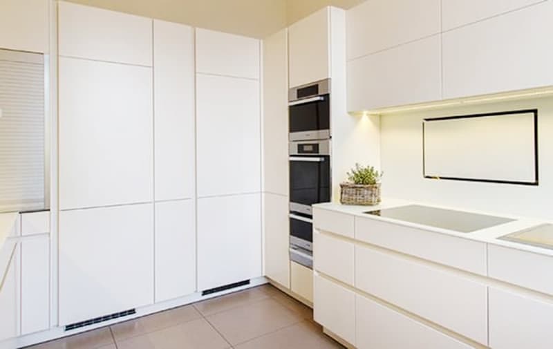 Modern, minimalist kitchen with white cabinets and drawers, built-in appliances including a dishwasher, and a large wall-mounted TV or projection screen.