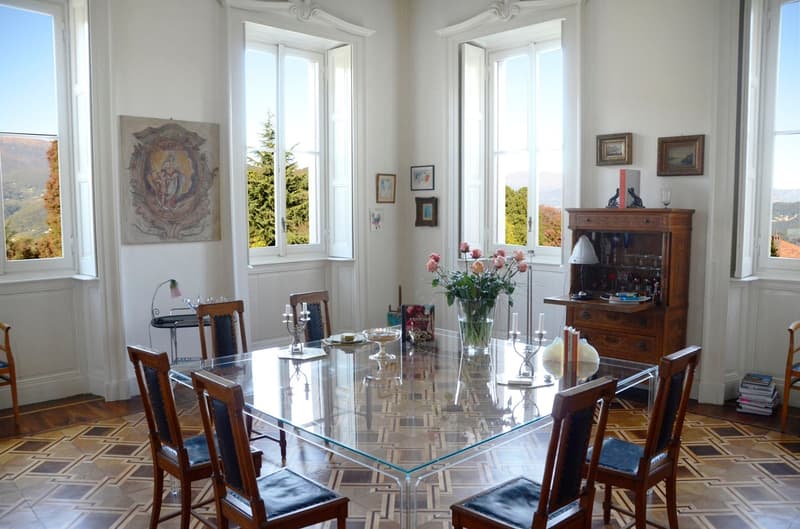 Spacious dining room with large windows offering a scenic view, ornate wall decor, wooden furniture including a glass-topped dining table and chairs, and various decorative items such as vases and framed artwork.