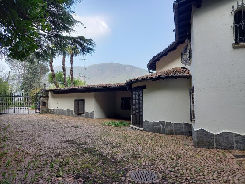 Two-story house, white walls, brown tiled roof, brick driveway, iron gate, black window shutters, fenced garden, mountainous landscape in the background