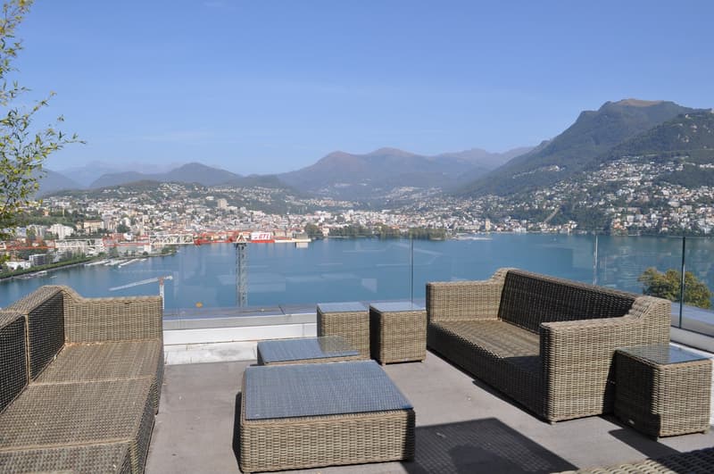 Outdoor terrace with wicker furniture overlooking a scenic lake surrounded by mountains and a city skyline in the background.