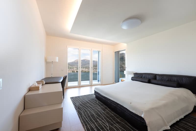 Spacious bedroom with large windows offering a scenic view of the mountains and lake, hardwood flooring, a king-size bed, and a modern nightstand.