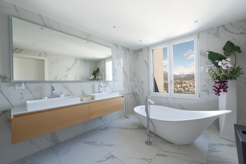 Spacious bathroom with marble-like tiles covering the walls and floor, a large freestanding bathtub, a double vanity with wooden cabinets, and a large window offering a scenic view of the city outside.