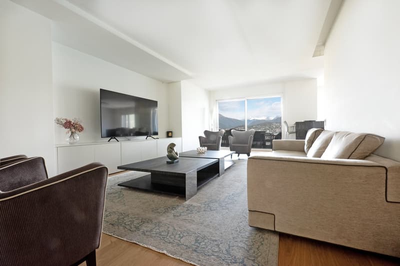 Spacious living room with large windows offering a scenic view, hardwood floors, beige sofa, black coffee table, TV mounted on the wall, and decorative elements such as a vase of flowers.