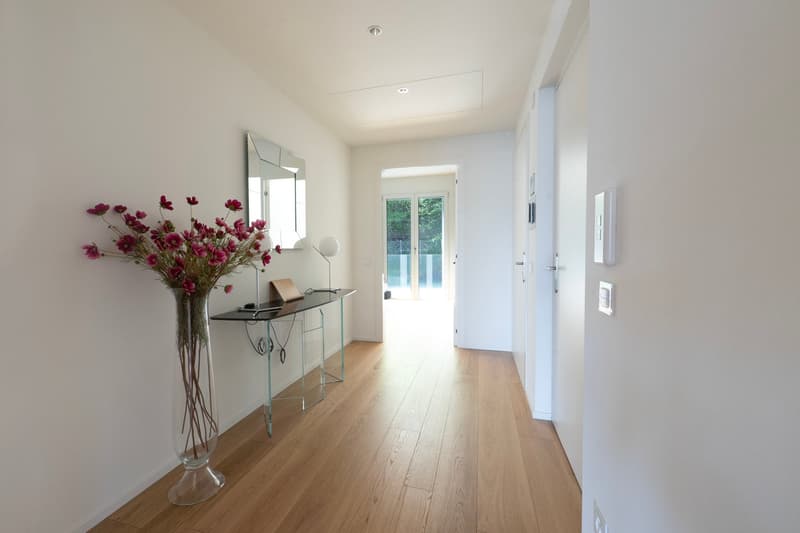 Bright and spacious hallway with hardwood floors, white walls, and a glass console table with a vase of pink flowers. The hallway leads to a door with a view of the outdoors.
