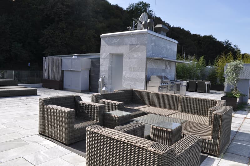 The image shows an outdoor living area with a stone structure that appears to have a fireplace and outdoor kitchen/grill. There are several wicker or rattan furniture pieces, including chairs and sofas, arranged in a seating area. The space is surrounded