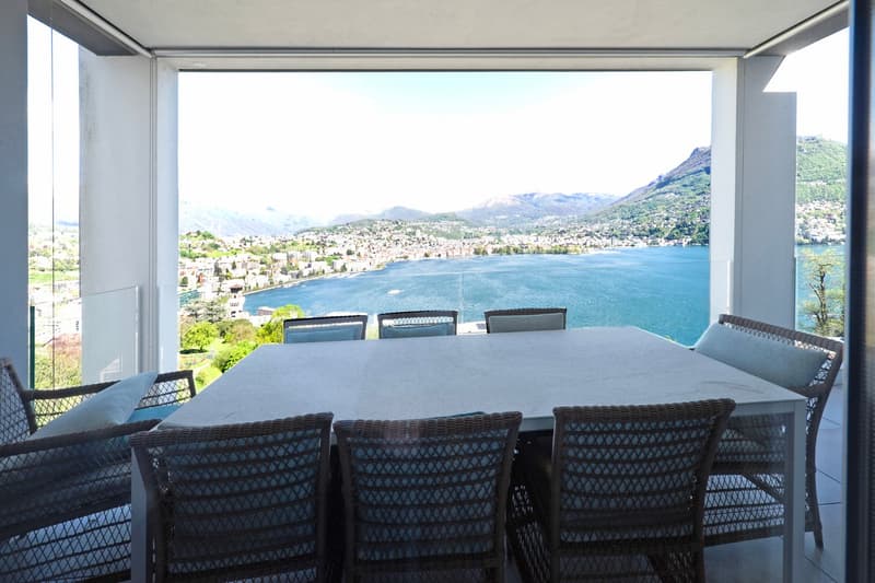 Large dining table with wicker chairs, overlooking a scenic lake and mountain landscape through large windows