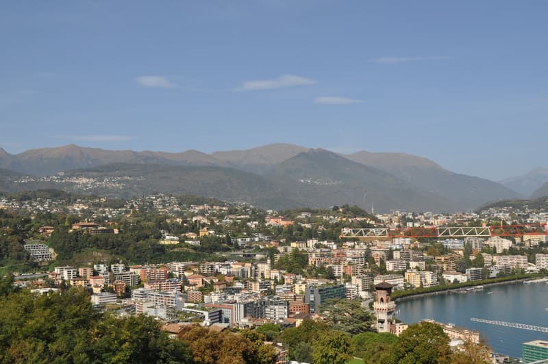 Panoramic view of a city surrounded by mountains, with a mix of residential and commercial buildings, bridges, and a body of water in the foreground.