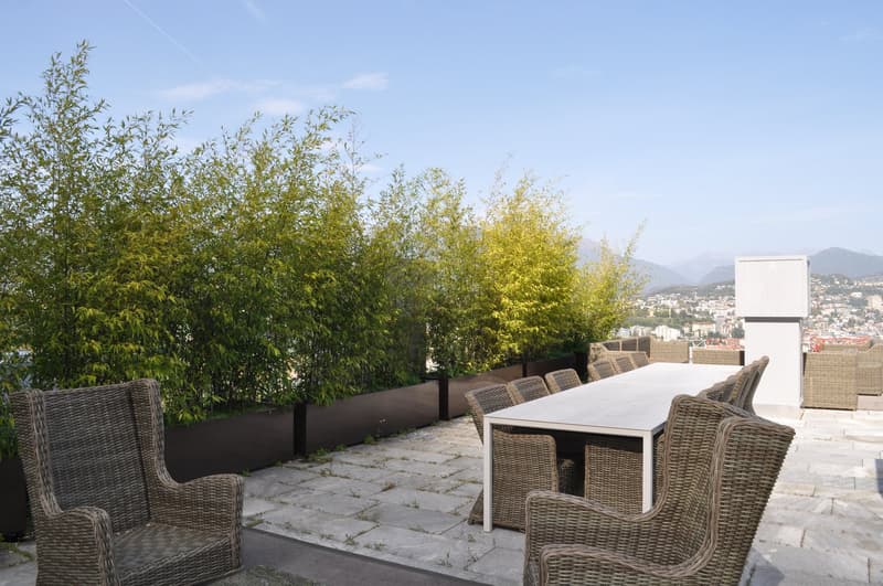 The image shows an outdoor patio or terrace area with a dining table and chairs made of wicker or rattan material. The patio is surrounded by lush greenery, including trees with yellow and green foliage. There is a white building or structure visible in t