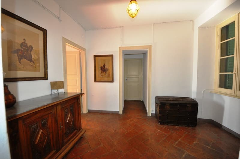 Spacious entryway with terracotta tile flooring, antique wooden furniture, framed artwork on the walls, and a decorative light fixture overhead. The room has a warm, rustic feel with a fireplace visible in the background.