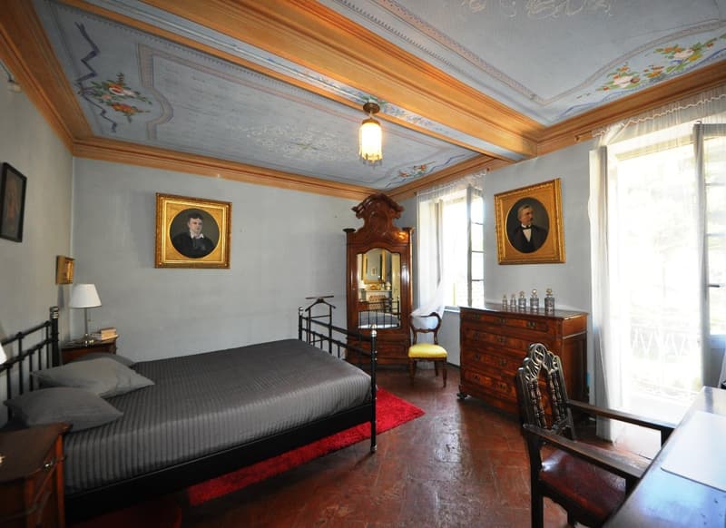 Large bedroom with ornate ceiling, wooden furniture including a dresser and armoire, framed portraits on the walls, and a fireplace.