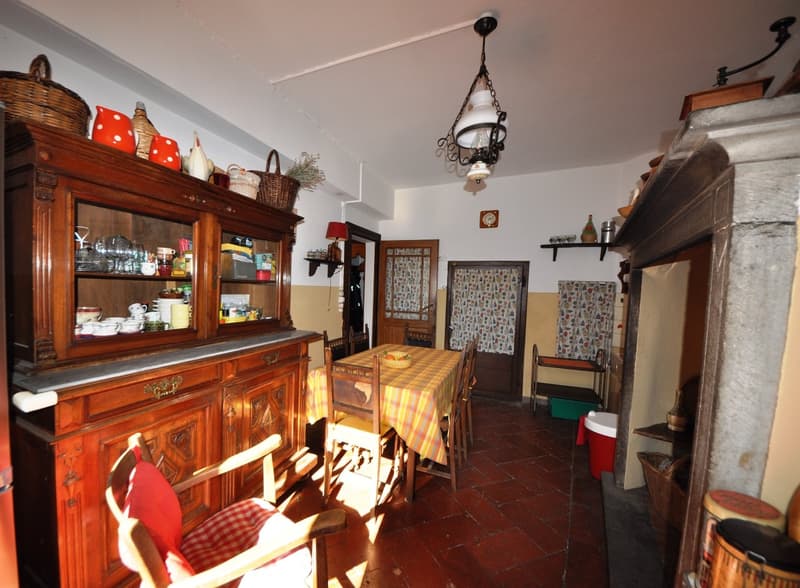 The image shows a cozy and rustic kitchen and dining area. The room features a large wooden cabinet with shelves and drawers, displaying various decorative items and kitchenware. There is a wooden dining table with chairs, and the walls are decorated with